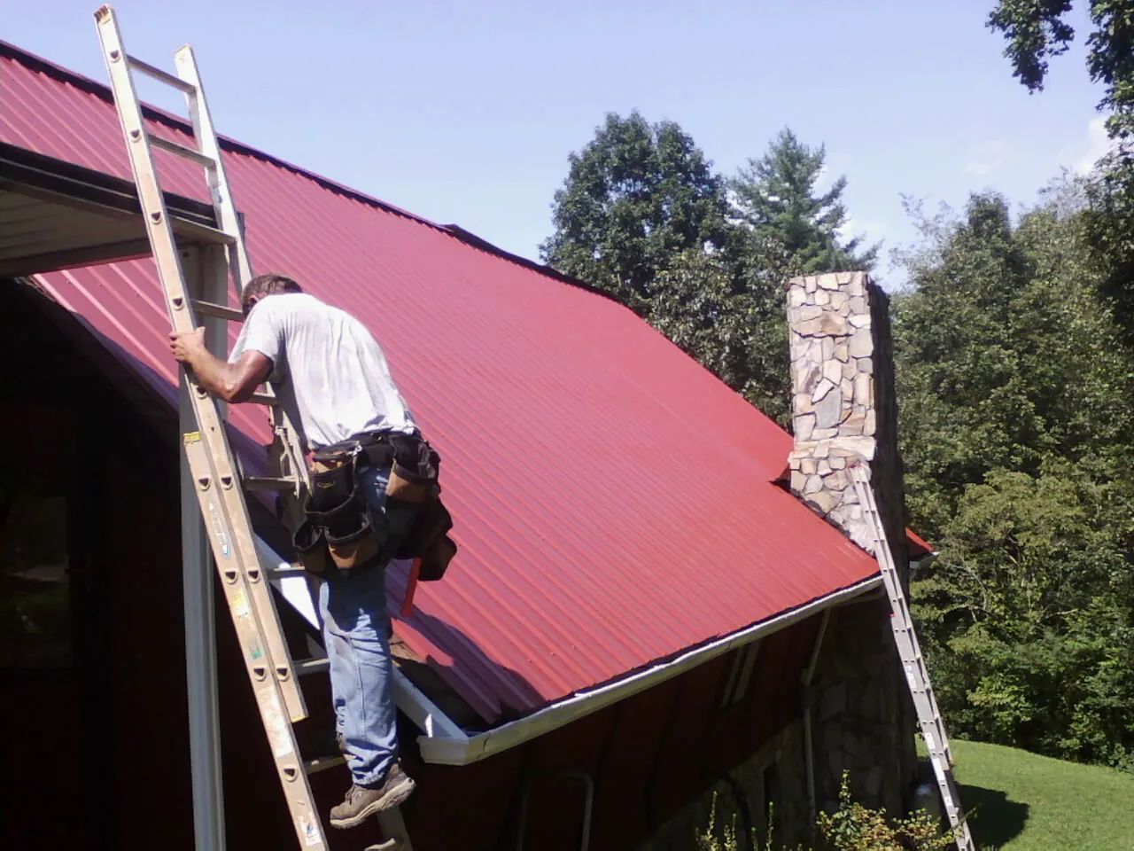 Roofer on a ladder beside a red metal roof and stone chimney — Soffit & Fascia Repair by Roofing Brothers in Lugoff